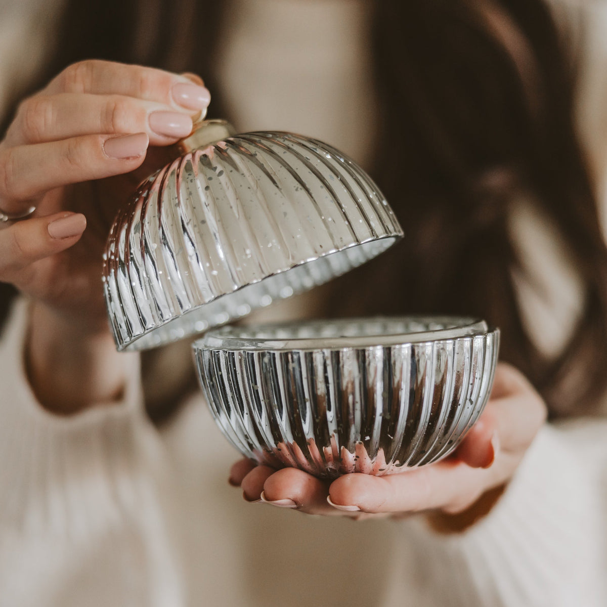 A person holds an open ribbed glass lidded container with a matching glass dome lid, shown in a close-up lifestyle shot against a soft beige sweater and blurred background.