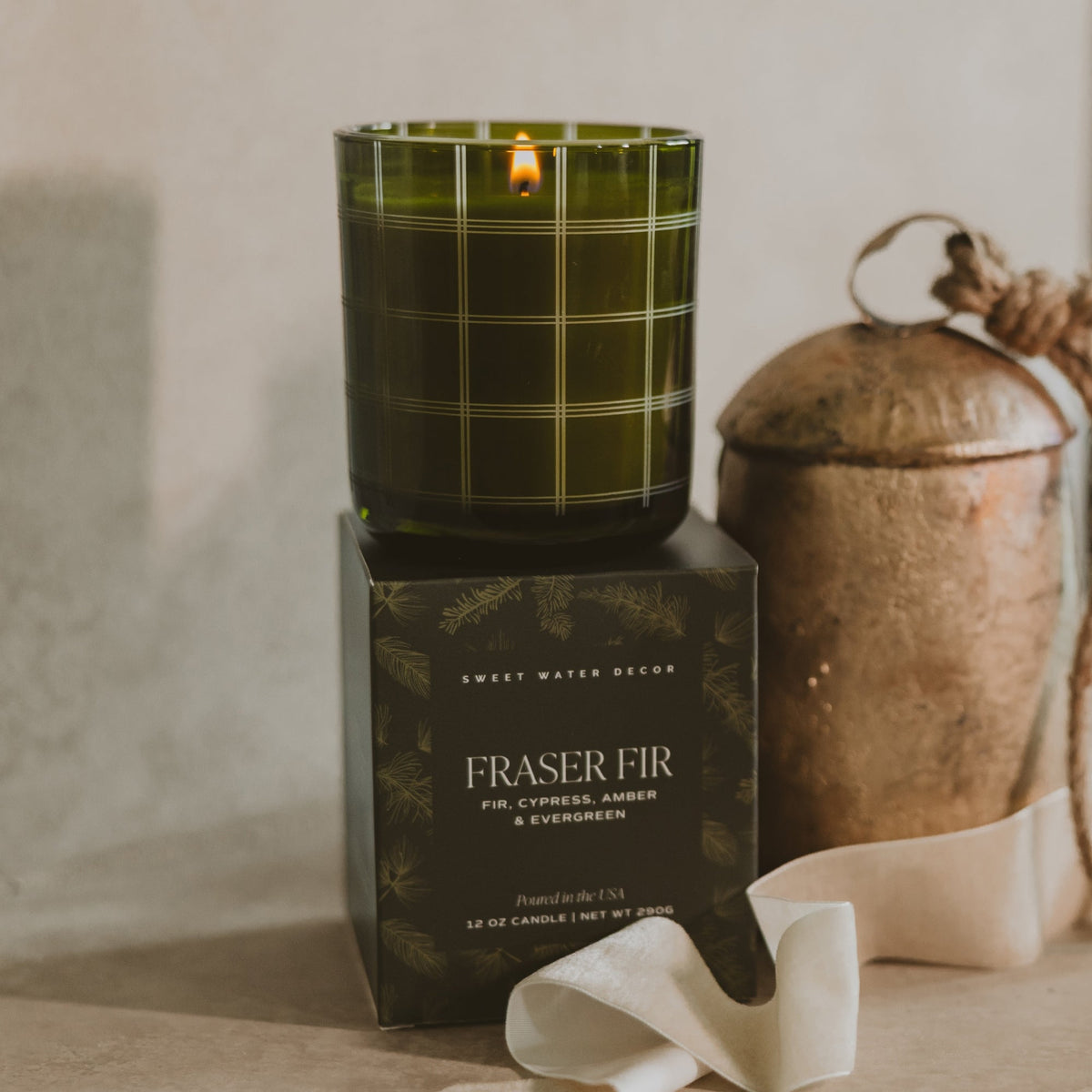 A lit candle in a dark green glass jar with a subtle plaid pattern sits on top of its black branded box. The box reads 'SWEET WATER DECOR' and 'FRASER FIR'.