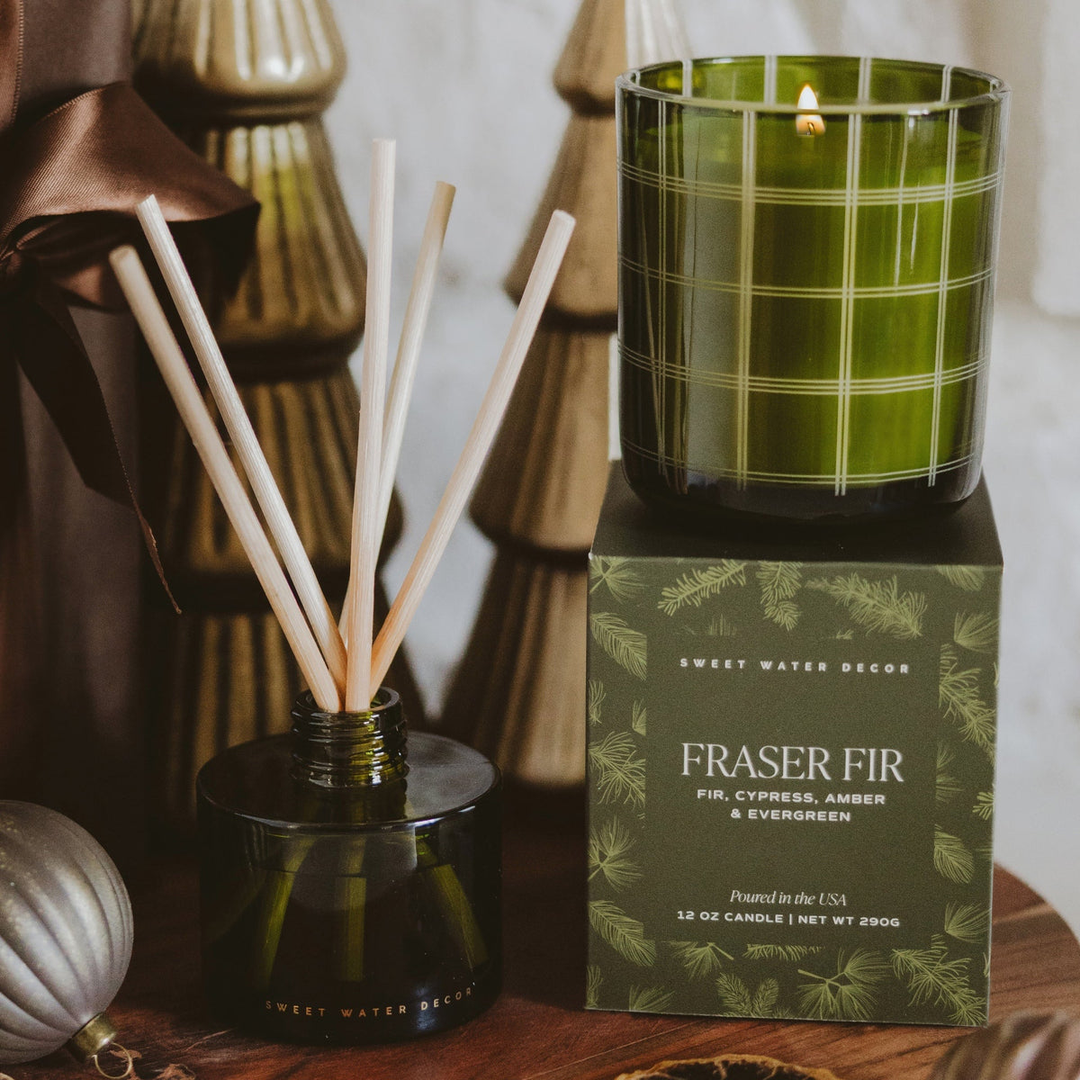 A green glass candle on top of its branded box next to a reed diffuser bottle with wooden reeds. The candle is in a translucent olive-green glass with a white windowpane plaid pattern, and the box reads 'FRASER FIR' by Sweet Water Scents.