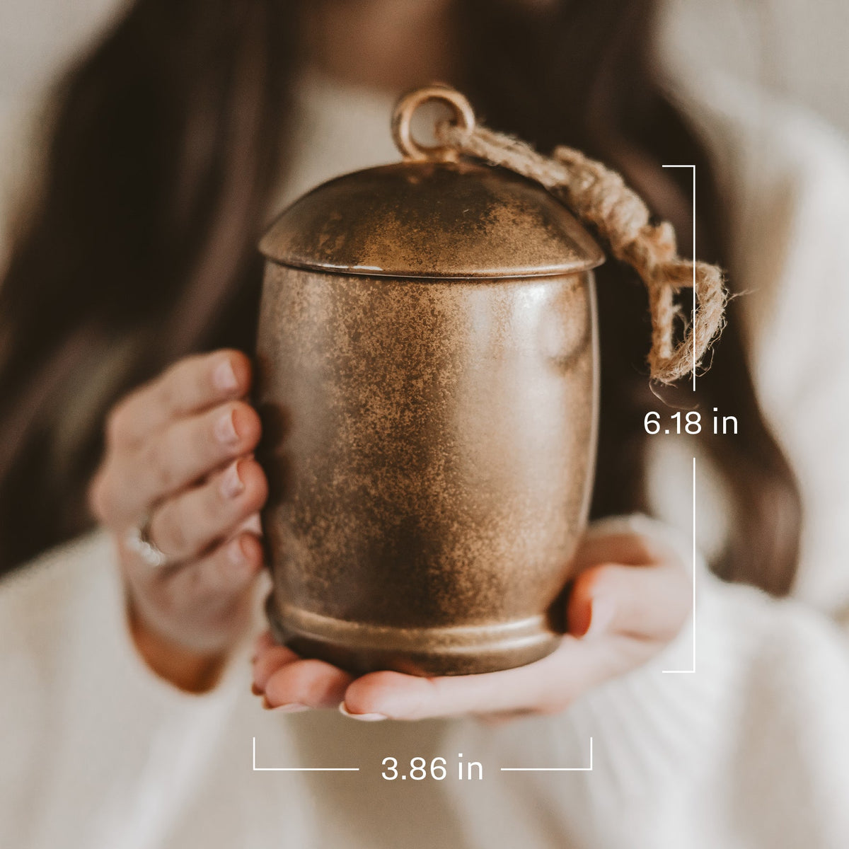 A person holds a small cylindrical metal keepsake box or urn with a lid and rope loop handle. White measurement lines and text indicate dimensions: 6.