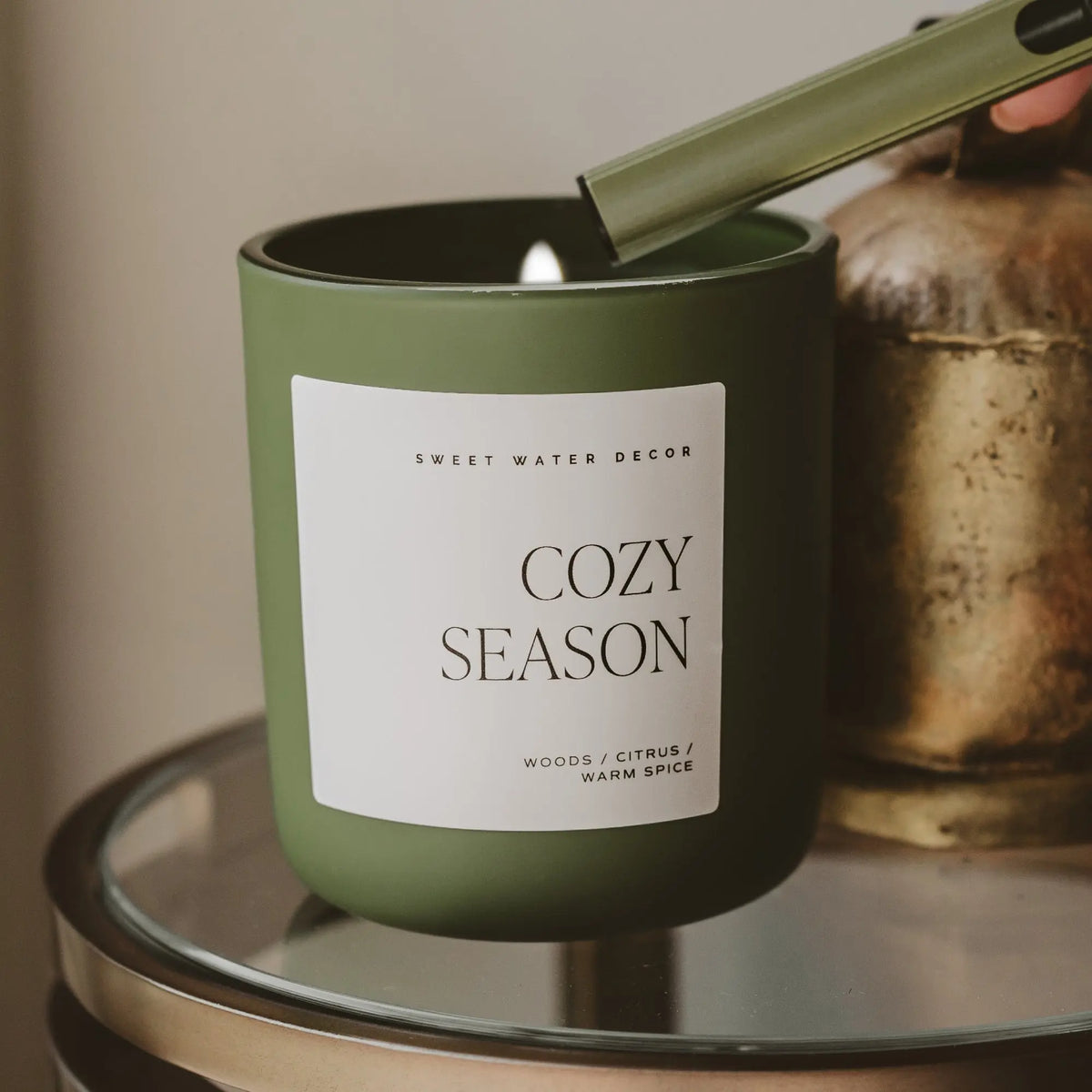 Green candle labeled 'Cozy Season' on a glass surface with a neutral background