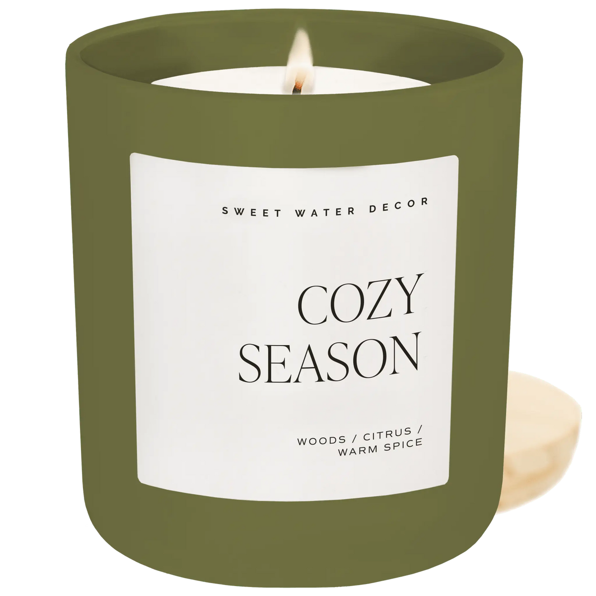 A lit candle in an olive green glass jar with a white label reading 'COZY SEASON' and 'SWEET WATER DECOR'. A small cream-colored candle is partially visible behind it on the right against a solid green background.