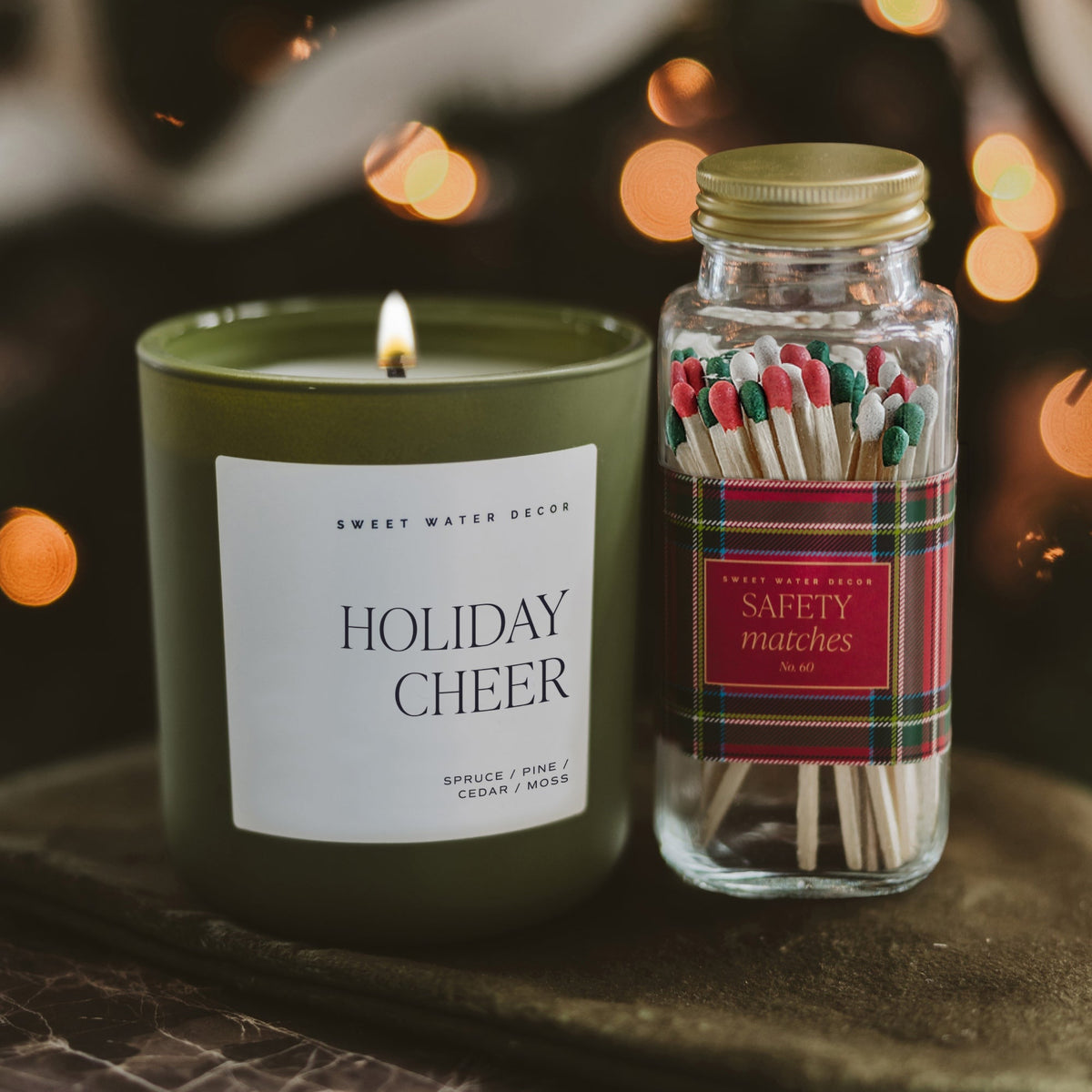 A lit green candle labeled 'Holiday Cheer' by Sweet Water Decor stands beside a clear glass jar of safety matches with a gold lid and a red plaid label. The scene is styled on a dark surface with warm blurred holiday lights in the background.