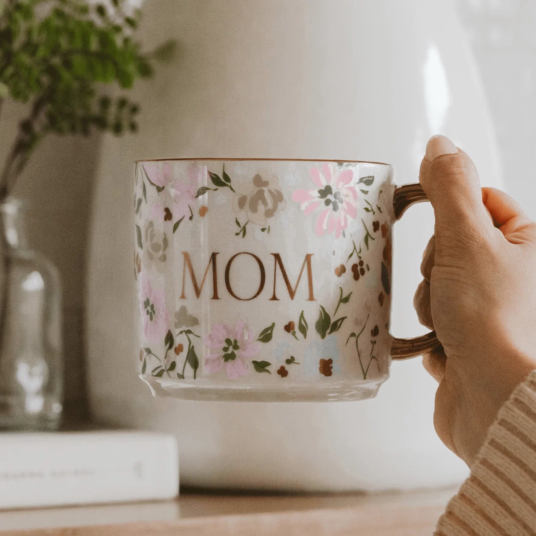 Hand holding a floral mug with 'MOM' printed on it, placed on a surface with a blurred background.