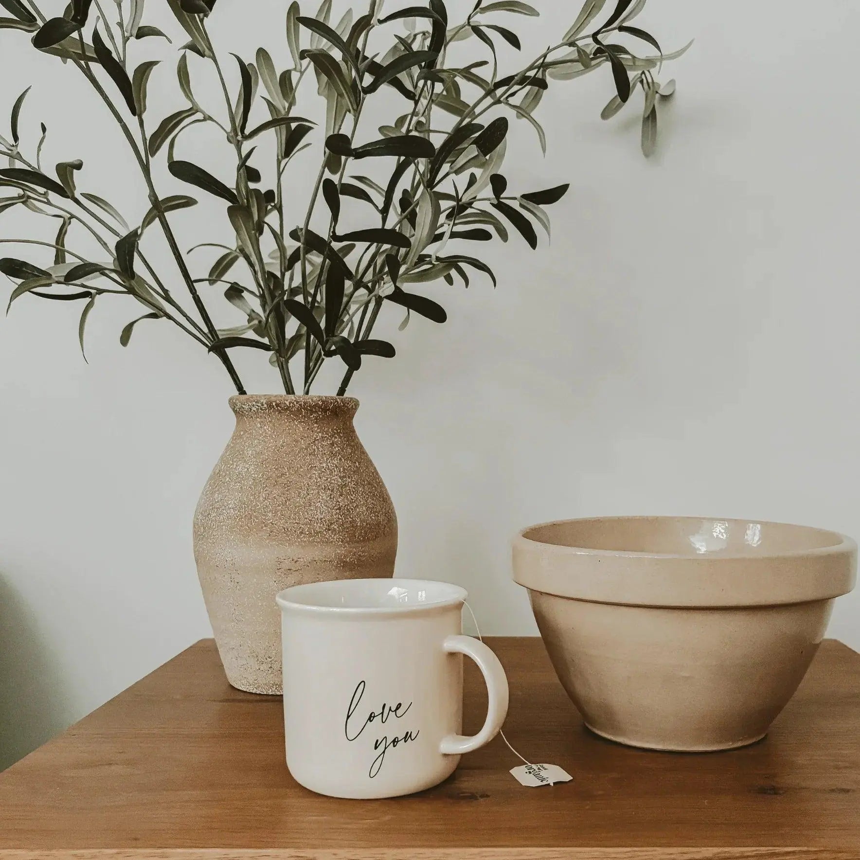 Cozy farmhouse decor with ceramic vase, olive branches, 'love you' mug, and stoneware bowl on wood table
