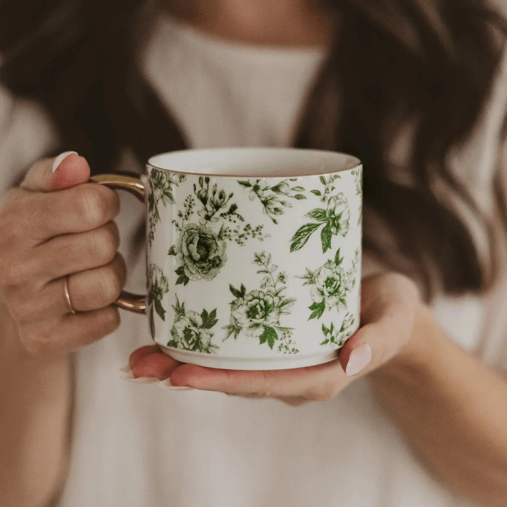 White ceramic mug with green floral pattern, gold handle, held by woman—cozy farmhouse decor