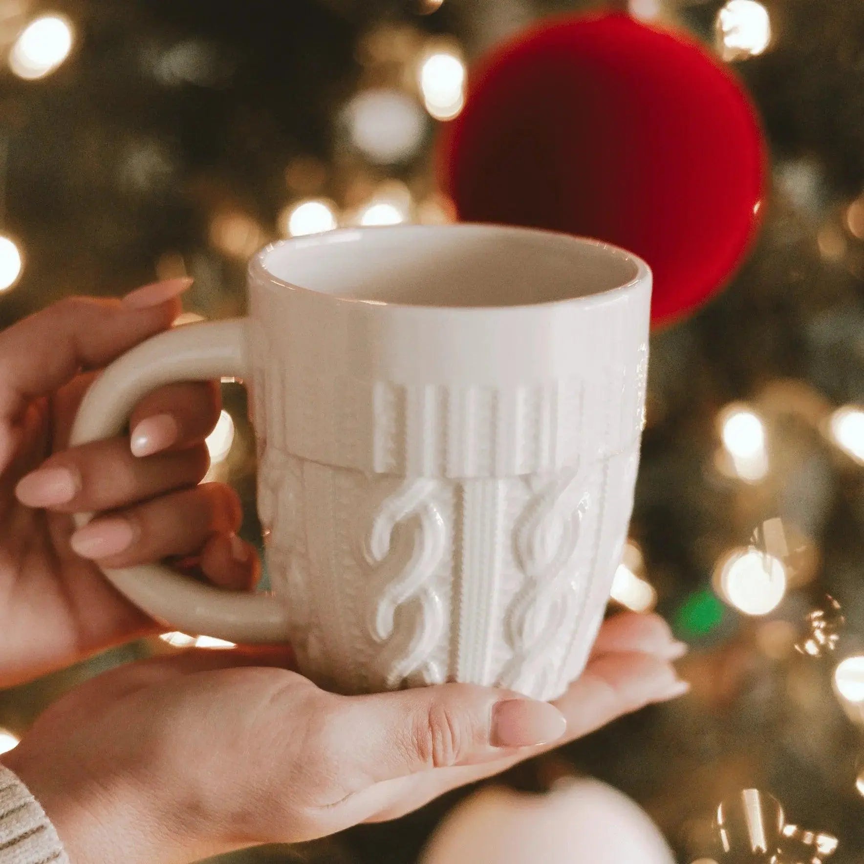 White cable knit mug held near Christmas tree lights, cozy farmhouse holiday decor