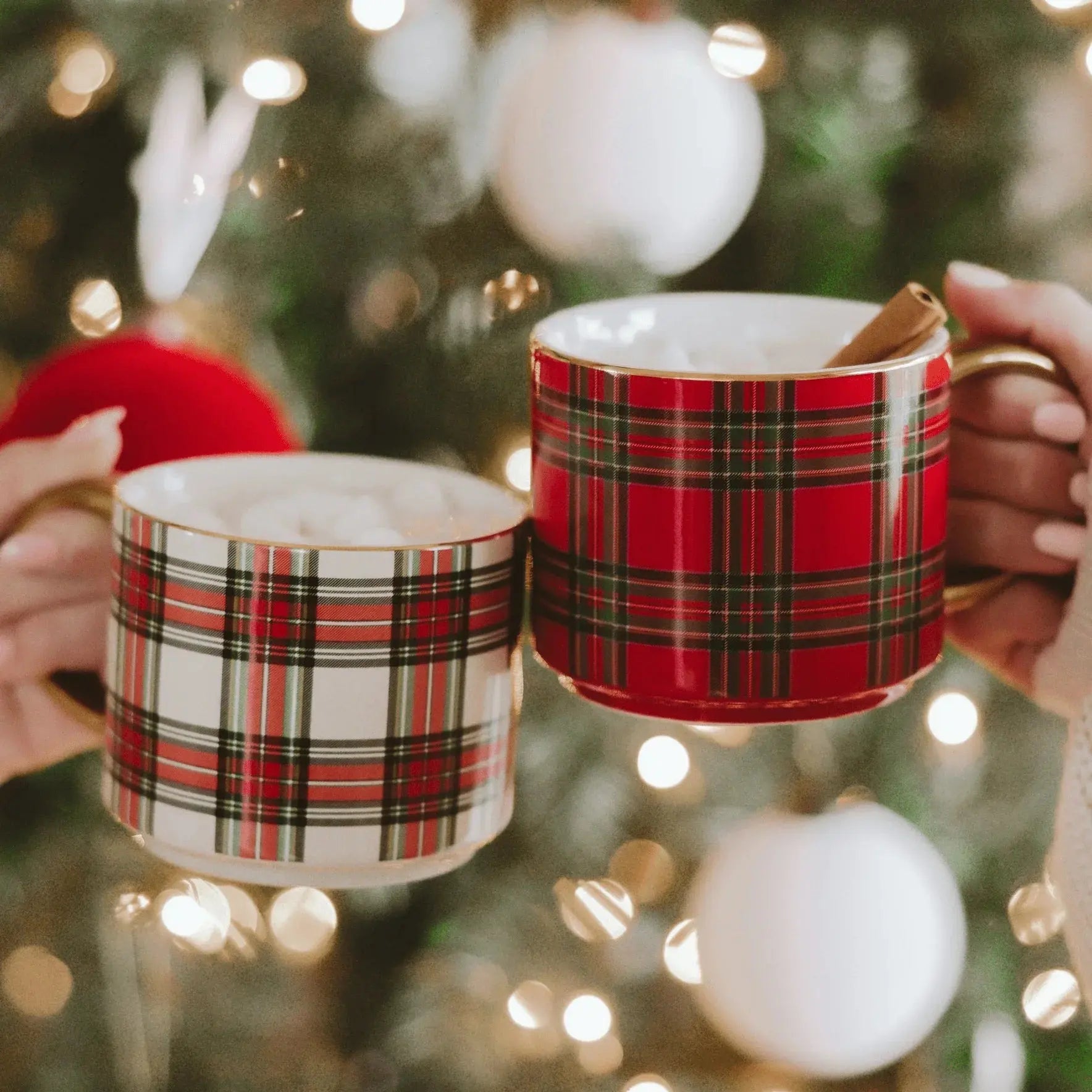 Red and white plaid mugs with gold rims, filled with hot cocoa and cinnamon sticks, held in front of a lit Christmas tree.