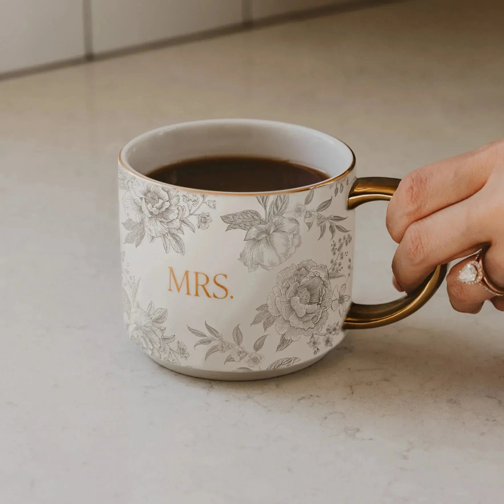 White floral mug with gold 'MRS.' text and handle, held on kitchen counter, cozy farmhouse decor