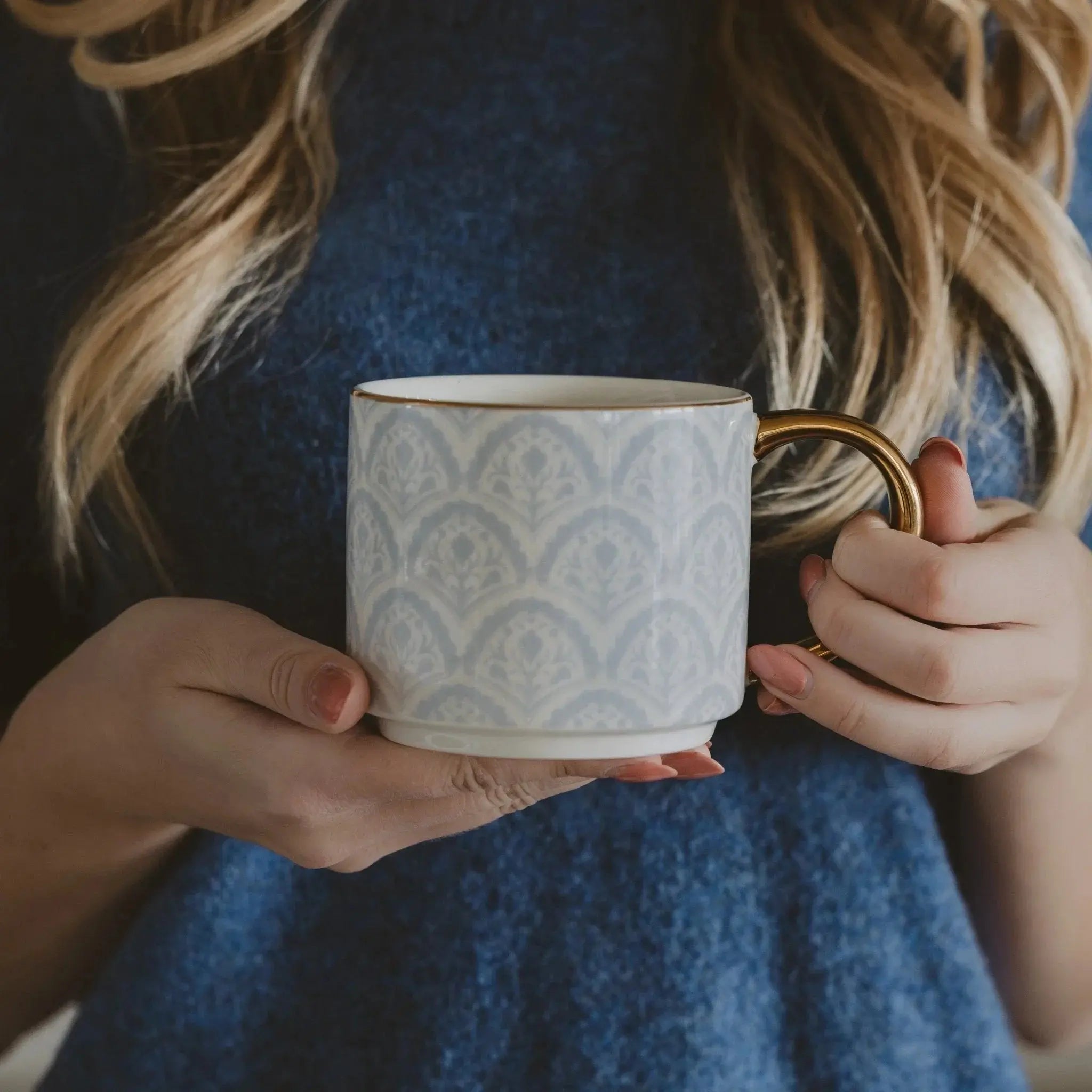 Woman holding elegant ceramic mug with gold handle and blue-gray damask pattern, cozy farmhouse style
