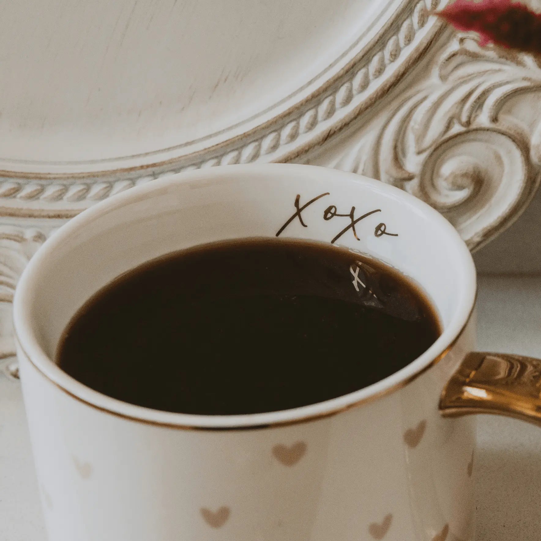 White mug with gold hearts and 'xoxo' text, filled with dark liquid, on a decorative surface.
