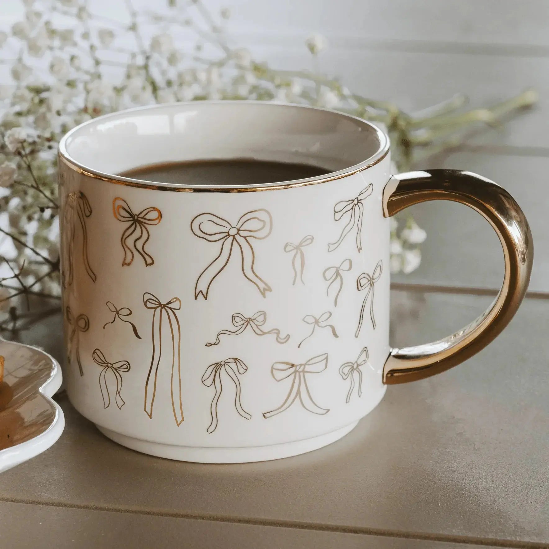 White ceramic mug with gold bow pattern and gold handle, filled with coffee, cozy farmhouse decor