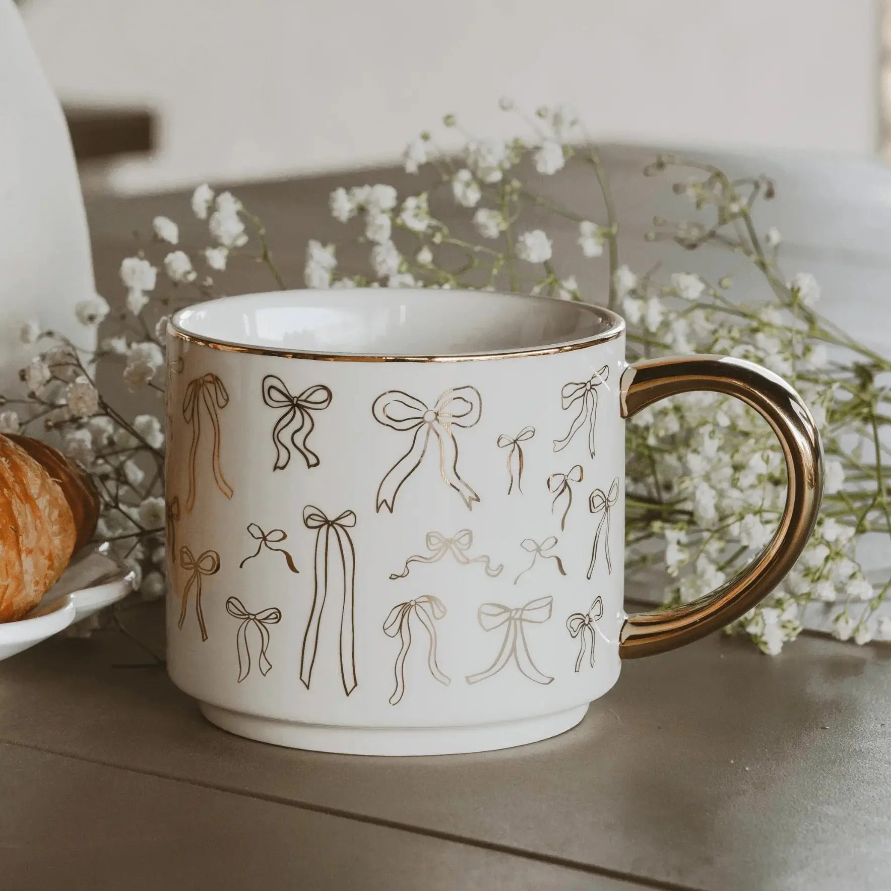 White ceramic mug with gold bow pattern and gold rim, cozy farmhouse decor with baby's breath flowers