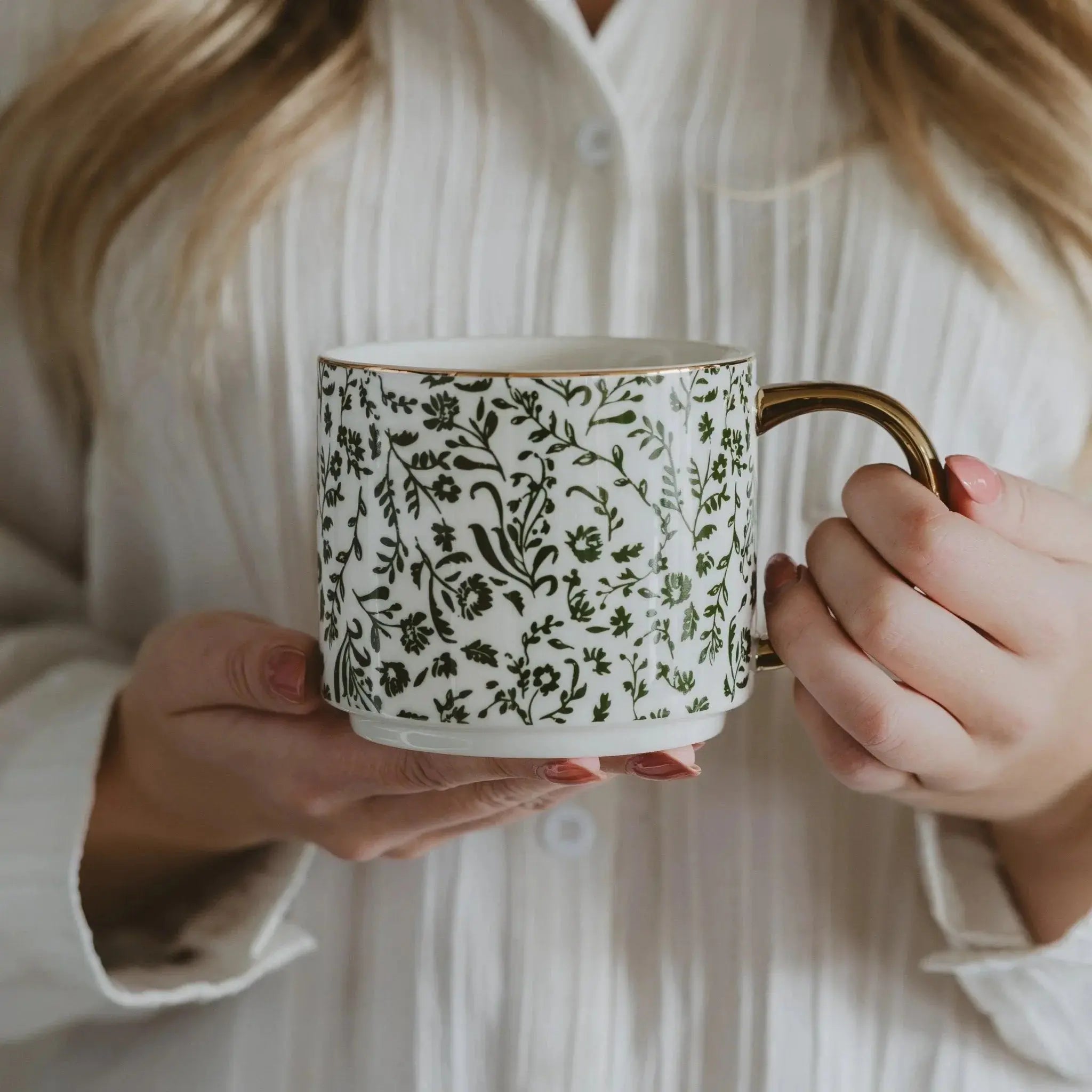 White ceramic mug with green floral pattern and gold handle, held by woman in cozy farmhouse style