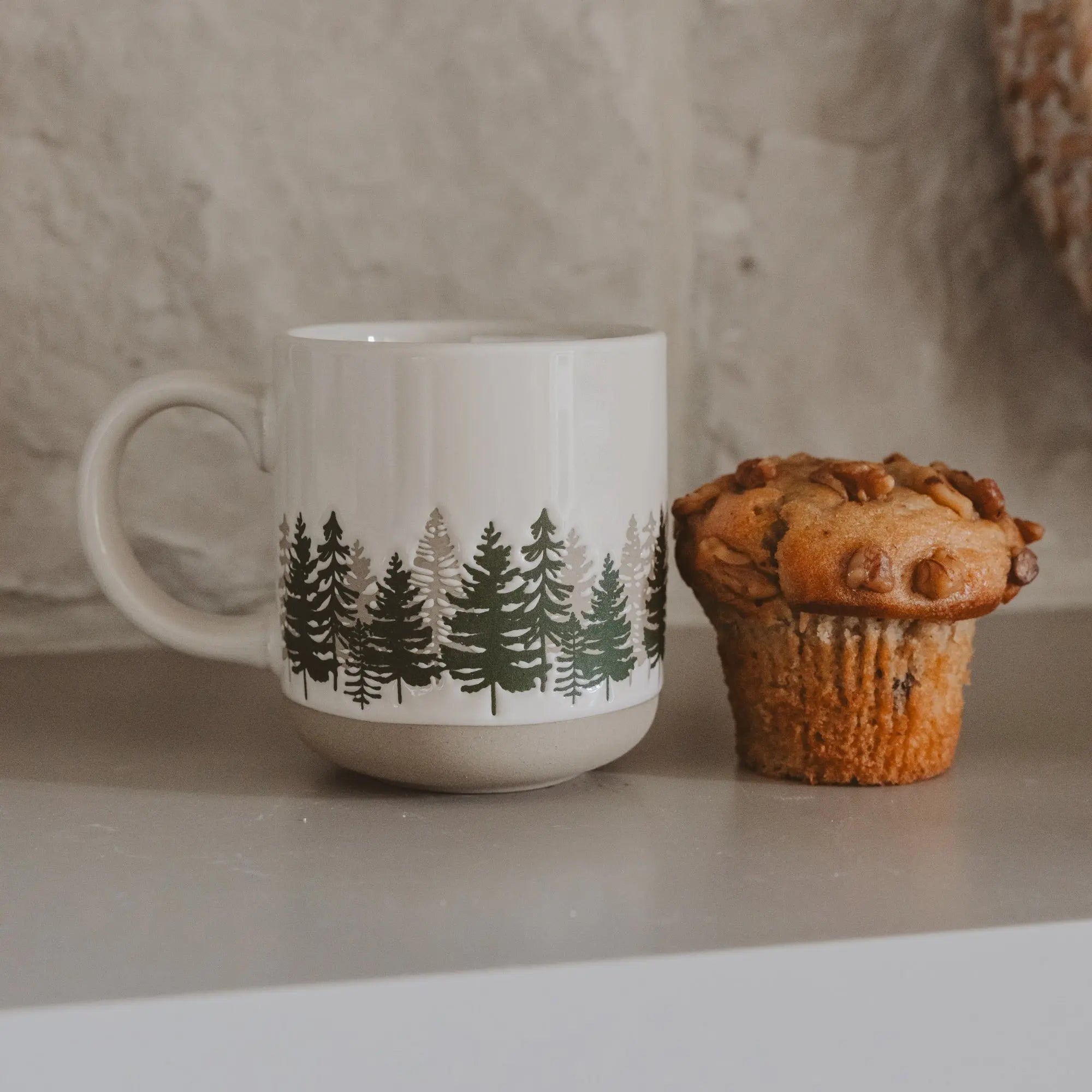 White mug with tree design next to a muffin on a neutral background