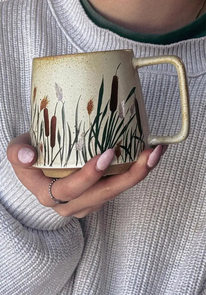 Hand holding rustic ceramic mug with cattail and reed design, cozy farmhouse style home decor