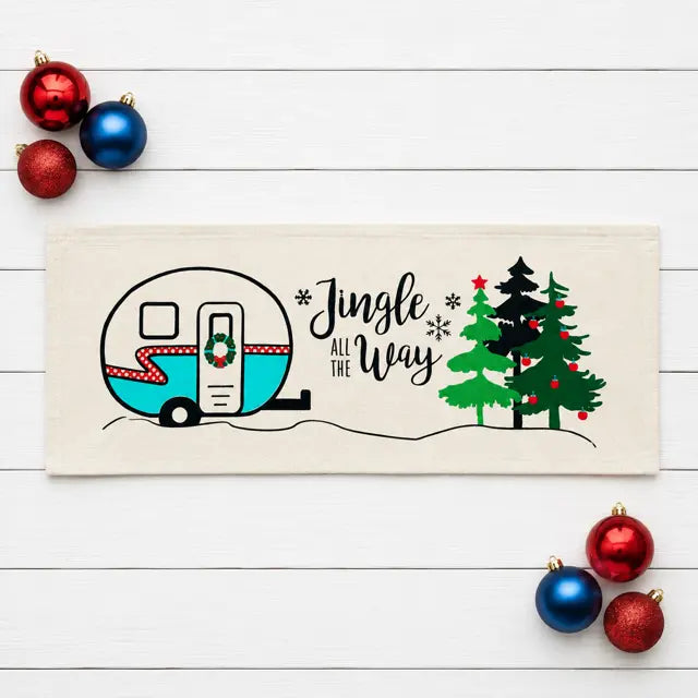 A rectangular cream-colored fabric tea towel or runner laid flat on a white wood background, decorated with a black line-art illustration of a retro camper, Christmas trees, snowflakes, and the phrase 'Jingle All The Way.' Red and blue ornaments are placed around it as styling props.