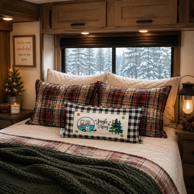 A cozy bedroom scene inside an RV or cabin, featuring a bed with plaid and buffalo check pillows, a decorative pillow with a camper illustration and the words 'Jingle a Ways,' a lit bedside lamp, and a snowy forest visible through the window.