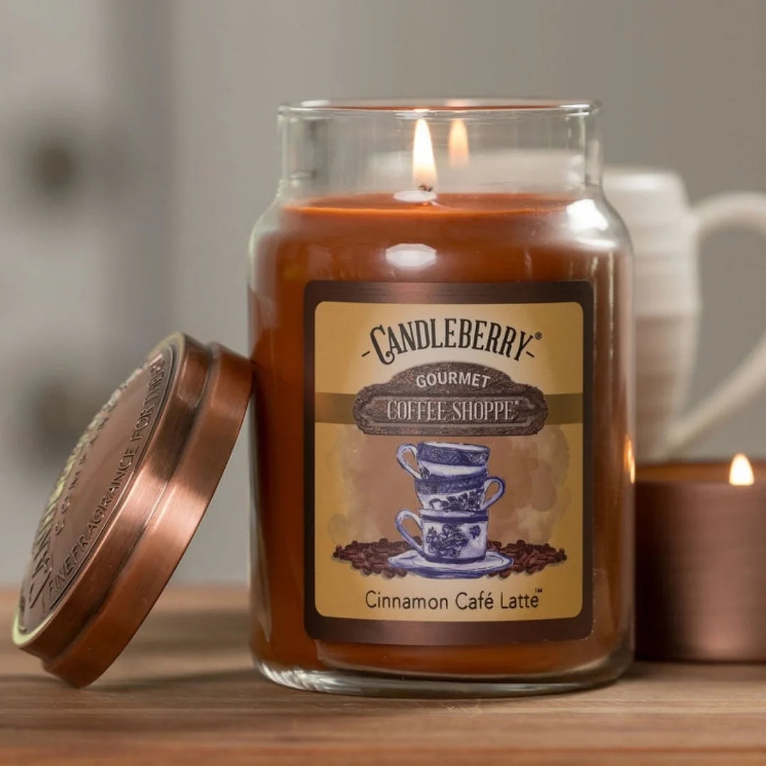 A lit Candleberry Gourmet Coffee Shoppe scented candle in a clear glass jar with a copper-toned lid leaning beside it on a wooden surface. The label reads Cinnamon Café Latte, and a blurred coffee mug and another lit candle appear in the background.