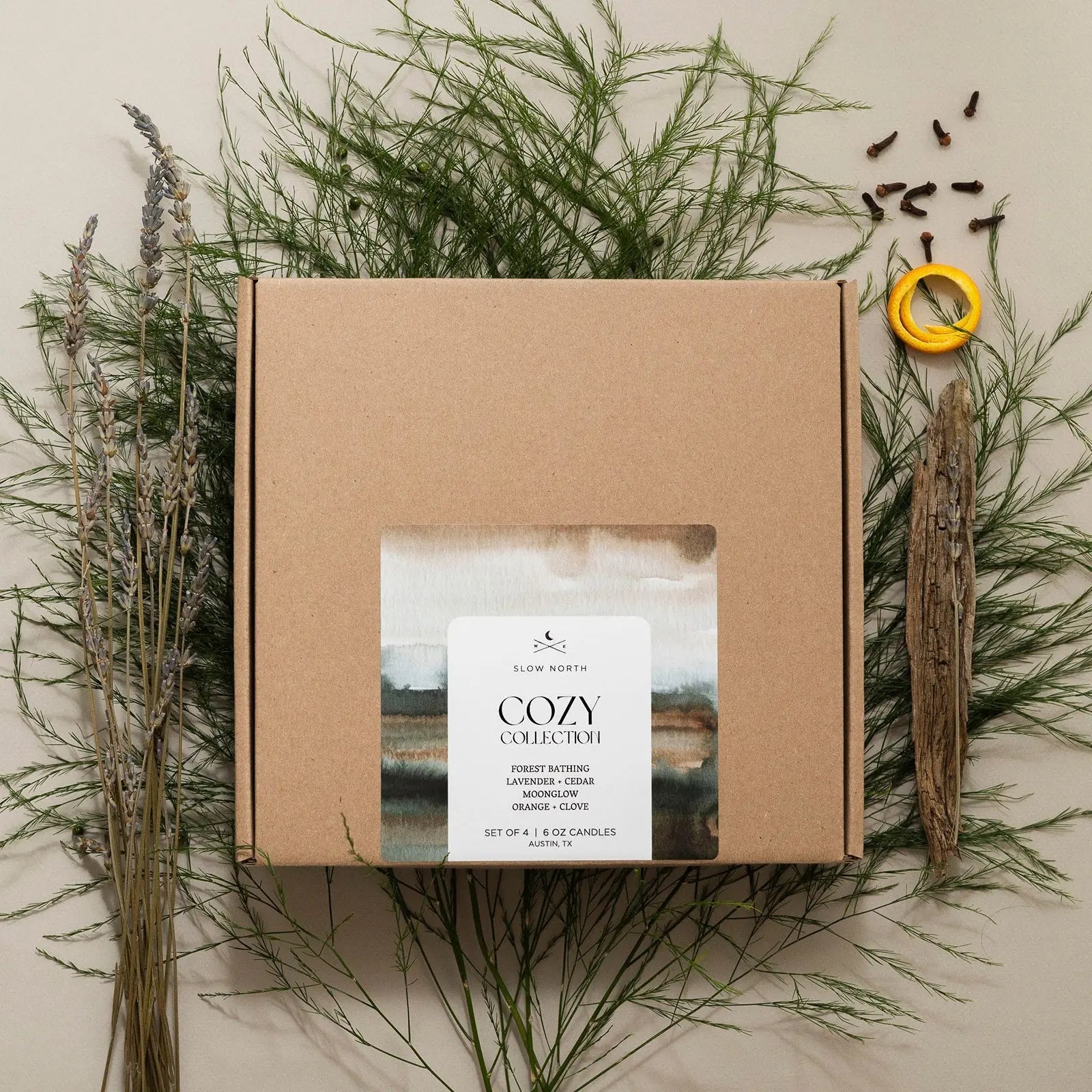 Cozy Collection candle set with lavender, cedar, orange, and clove scents, nestled in dried greenery and herbs.