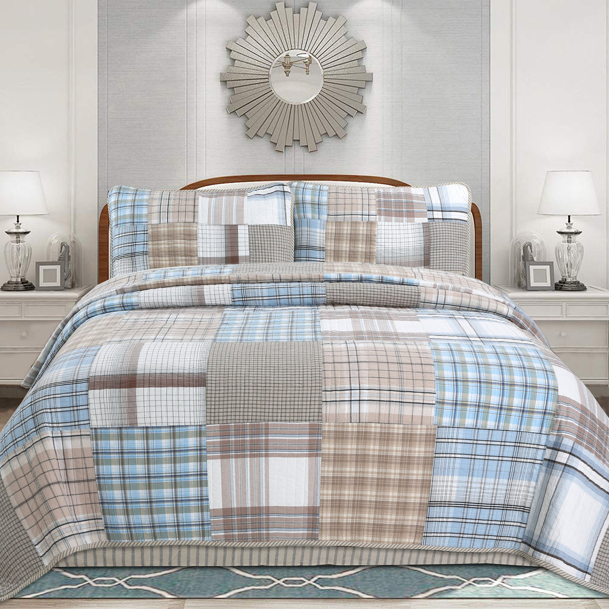 A bed dressed in a patchwork plaid quilt and matching pillow shams in shades of blue, beige, gray, and brown, styled in a symmetrical bedroom setting with matching nightstands, lamps, and a sunburst wall mirror.