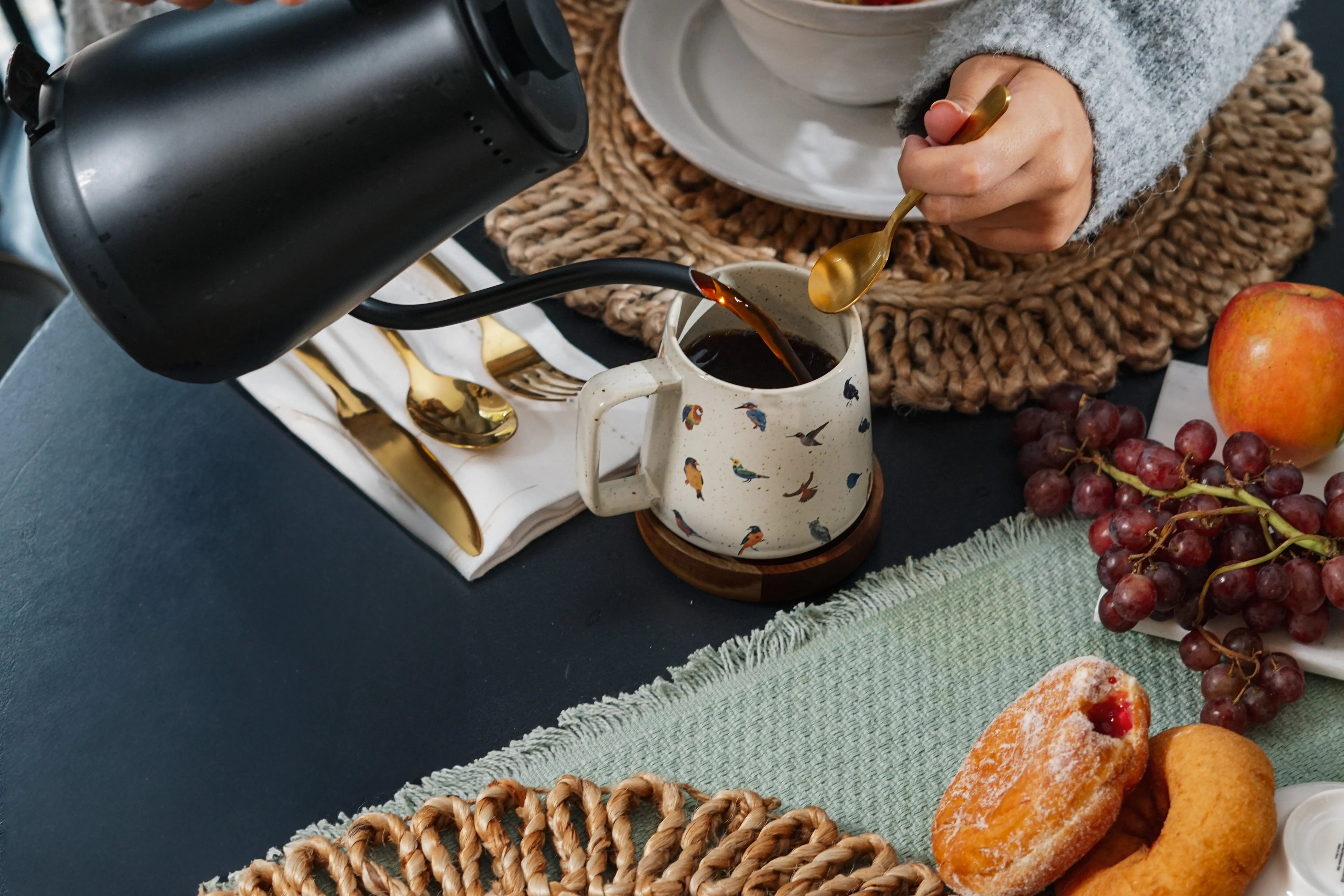 Cozy farmhouse breakfast with coffee poured into bird-print mug, fresh grapes, apple, pastries, and woven placemats