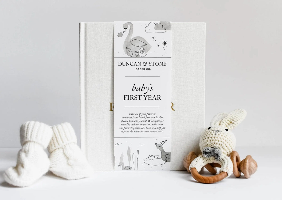 Book titled 'Baby's First Year' by Duncan & Stone with baby socks and a plush toy on a light gray background