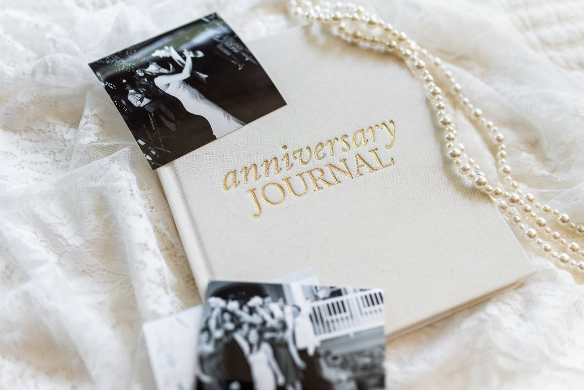 The Ivory Anniversary Journal with photos scattered on a cream cloth with pearls