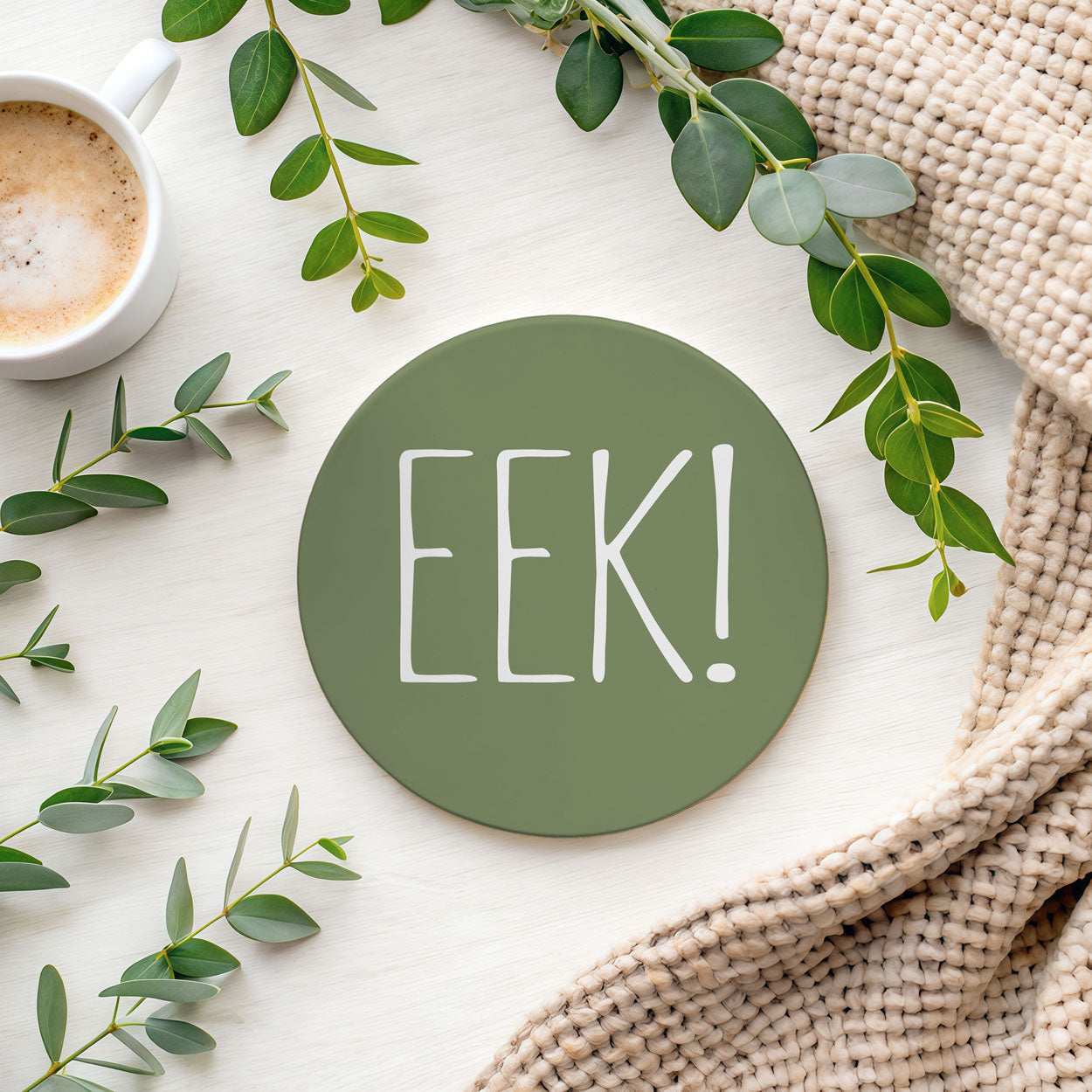Green round coaster with 'EEK!' text on a white surface with green leaves and a cup of coffee.