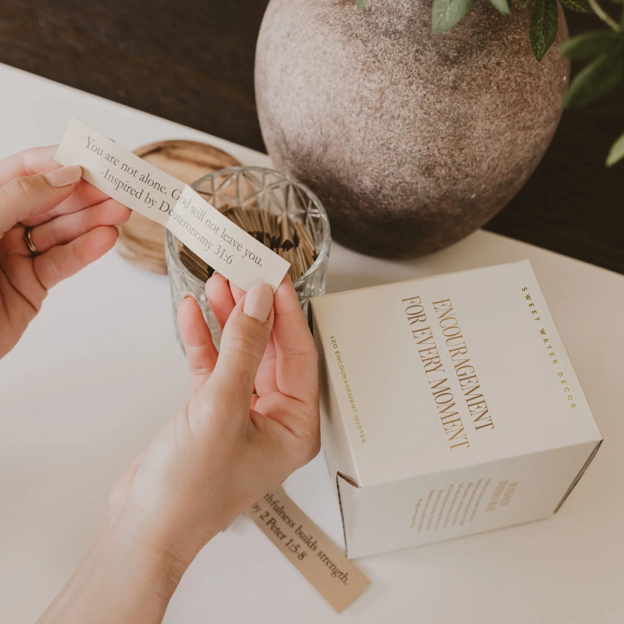 A person holds a small printed card in both hands beside a white box labeled 'FOREVER A MOMENT' and a glass jar, in a softly styled tabletop setting.