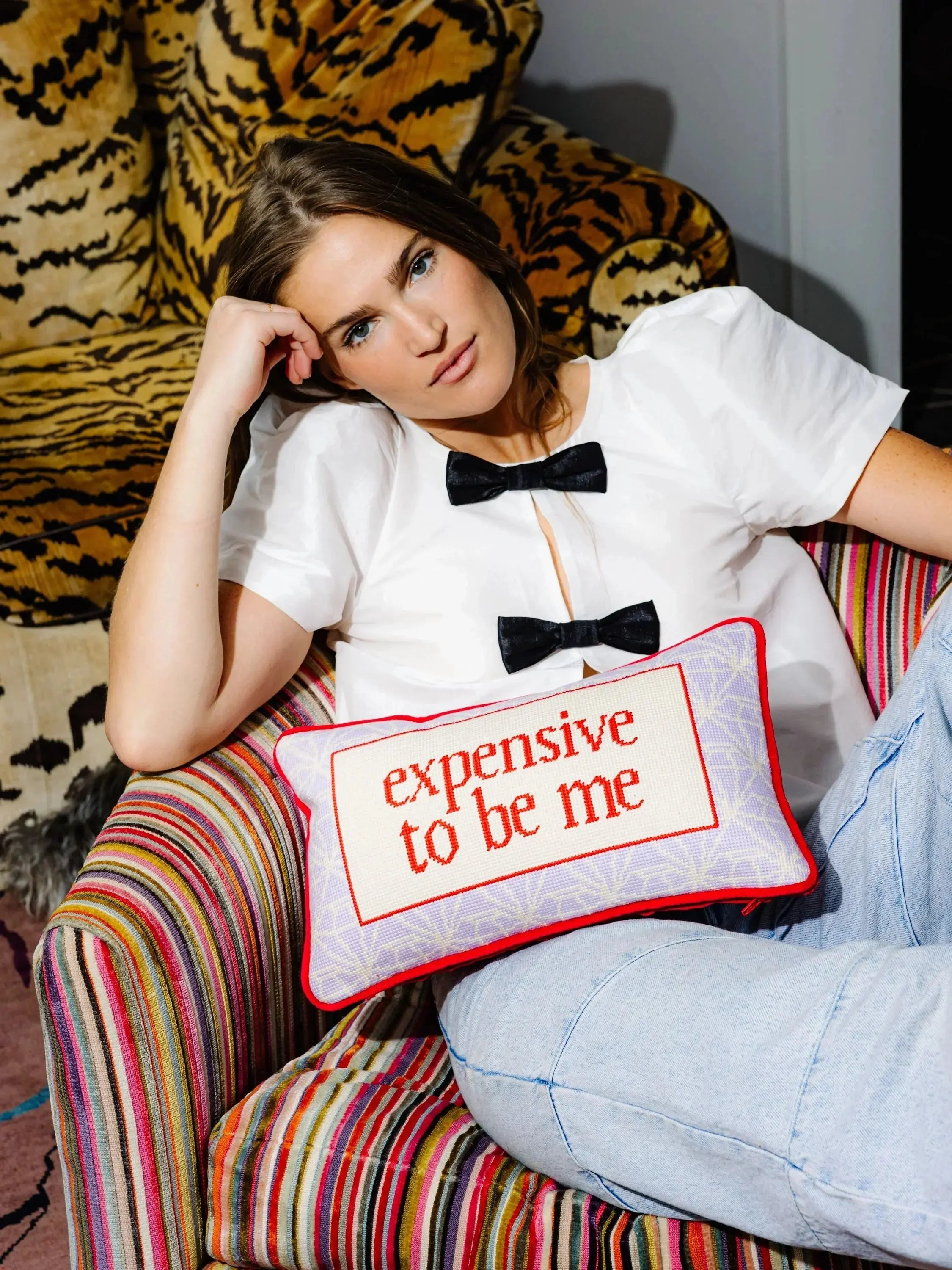 Woman sitting on a colorful chair with a pillow that says 'expensive to be me' in front of a tiger-patterned curtain.