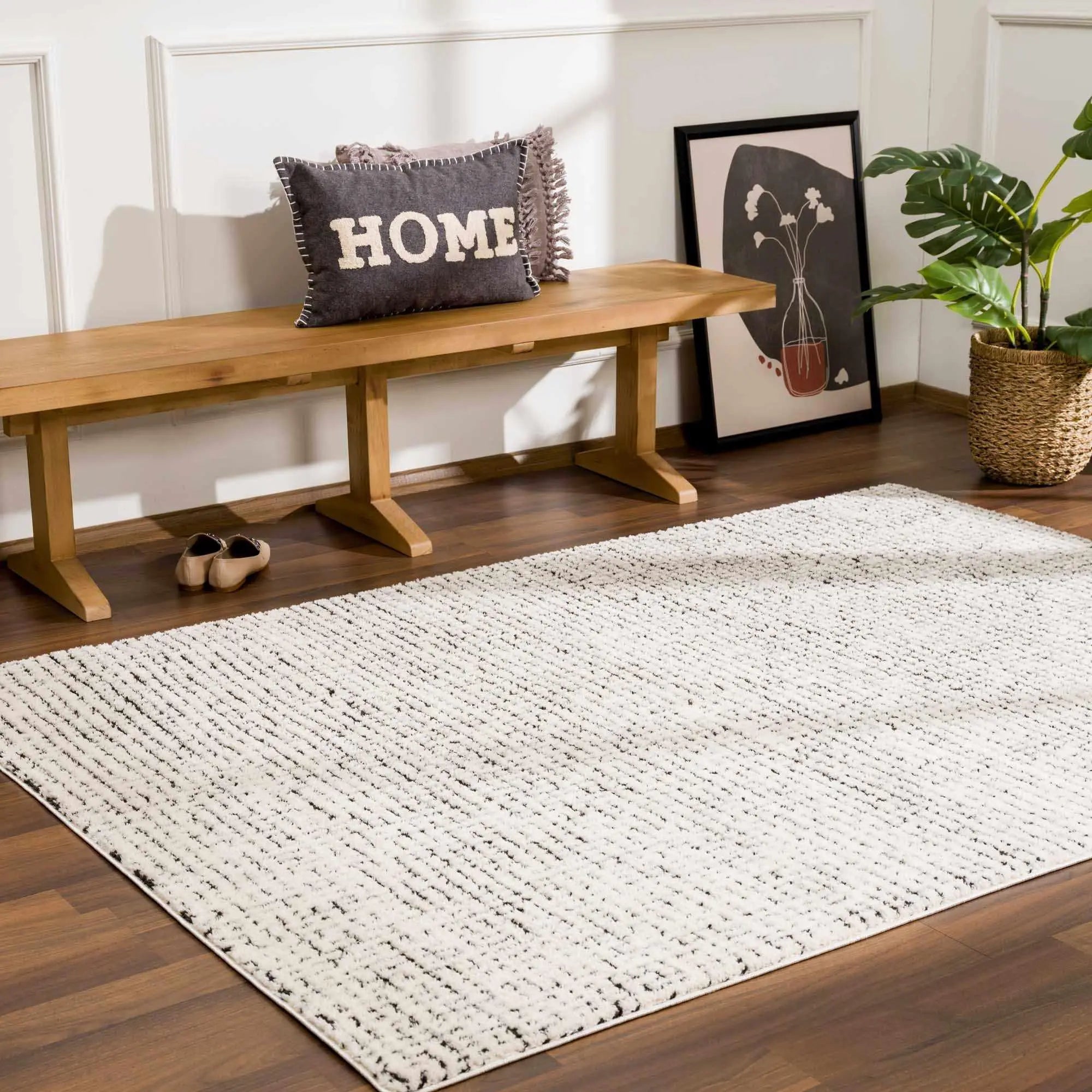 Cozy farmhouse area rug with textured white and black pattern, styled with wooden bench, 'HOME' pillow, and potted plant.