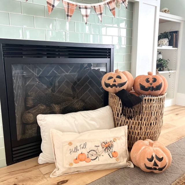 A styled fall-themed living room scene featuring a decorative cream pillow with a bicycle and pumpkins and the words 'Halloween Fun' in front of a fireplace, alongside two carved pumpkins in a wicker basket and another pumpkin on the floor.