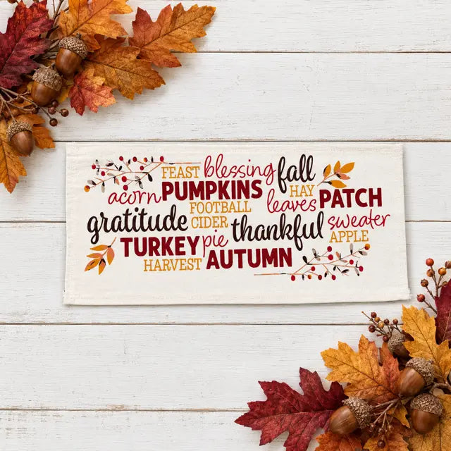 A rectangular cream fabric sign with autumn-themed typography and small leaf/berry illustrations, displayed on a white wood surface with fall leaves, acorns, and berries around it.