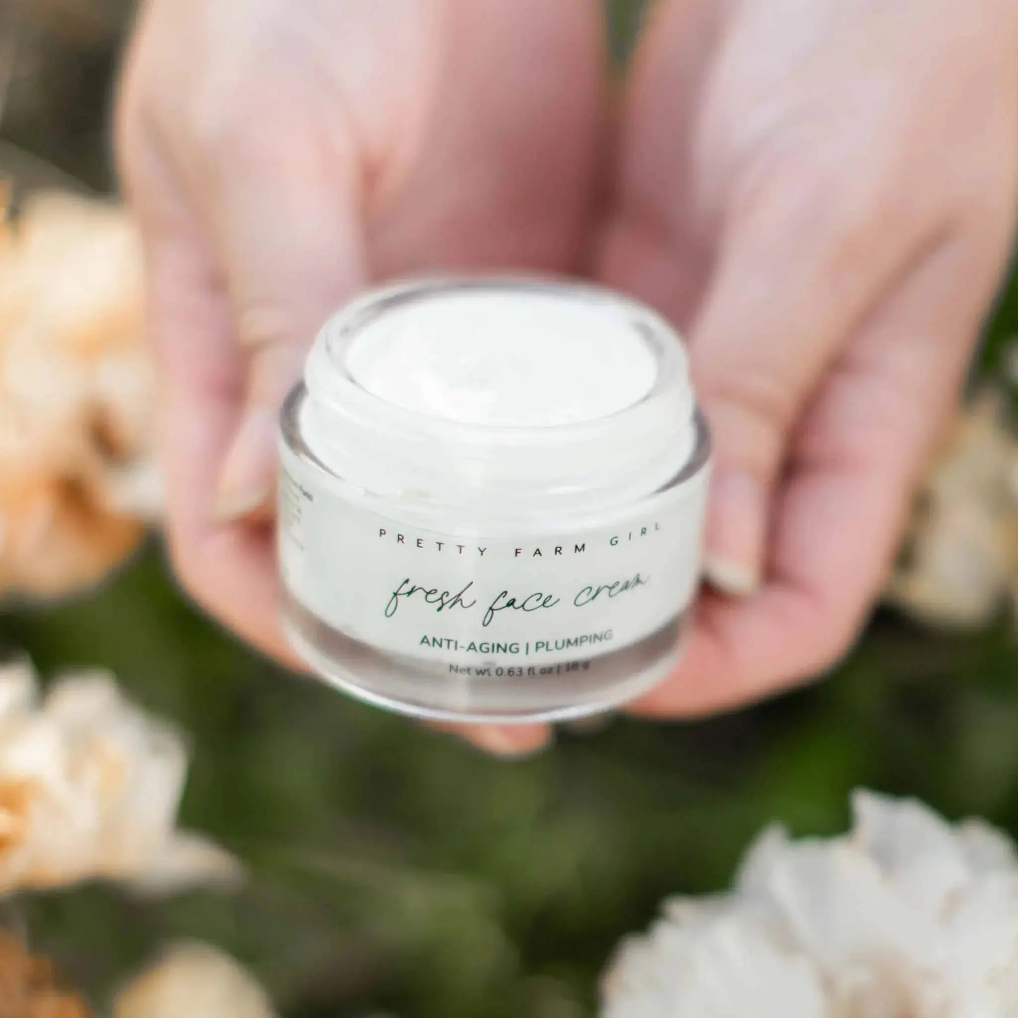 Pretty Farm Girl fresh face cream, anti-aging plumping moisturizer in clear jar held gently with floral background