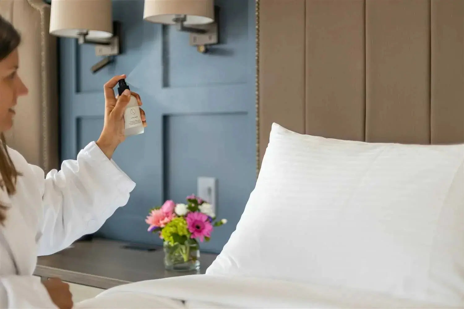 Woman spraying linen spray on cozy bedroom pillow, farmhouse decor with fresh flowers and soft lighting