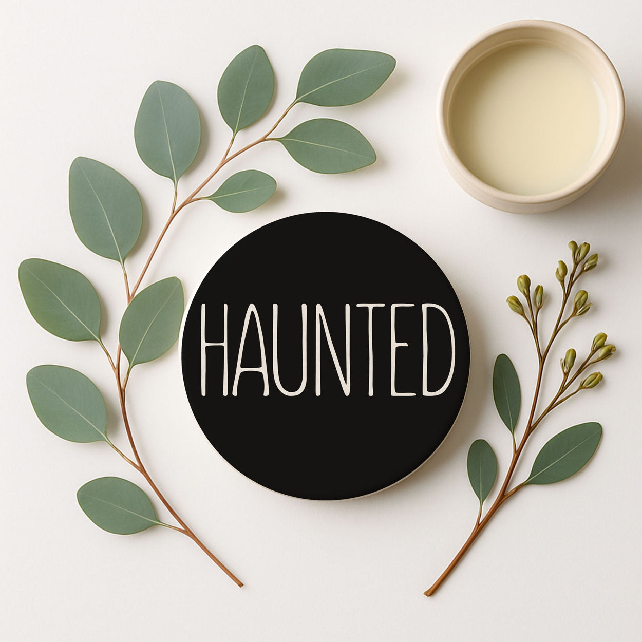 Black coaster with 'HAUNTED' text surrounded by green leaves on a white background
