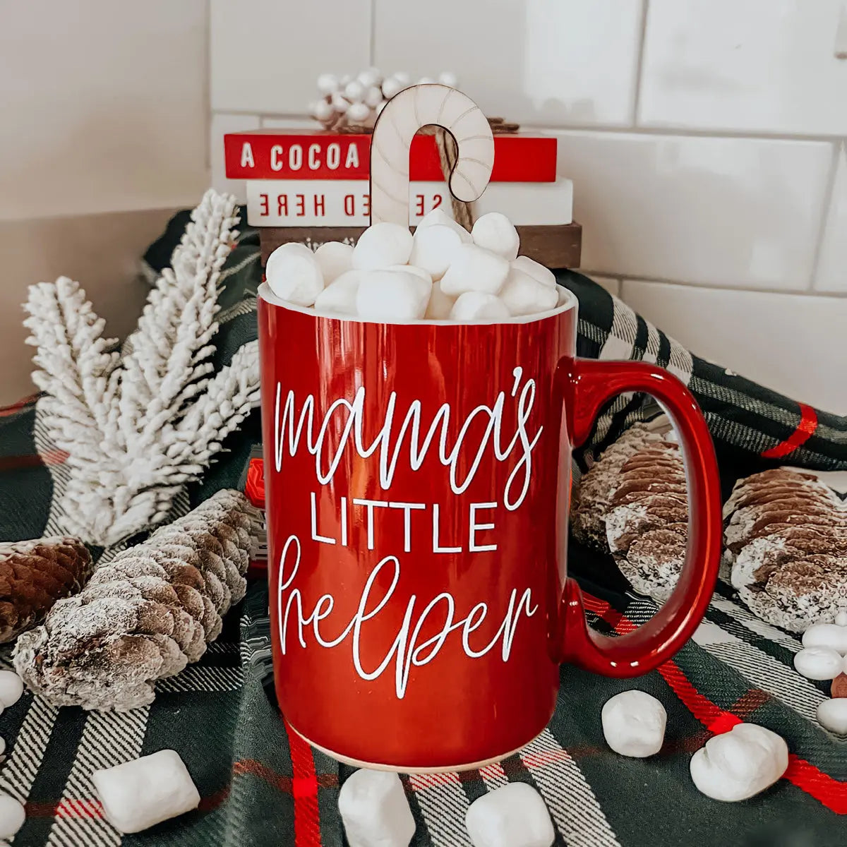 Red mug with 'mama's little helper' text filled with marshmallows on a festive background.