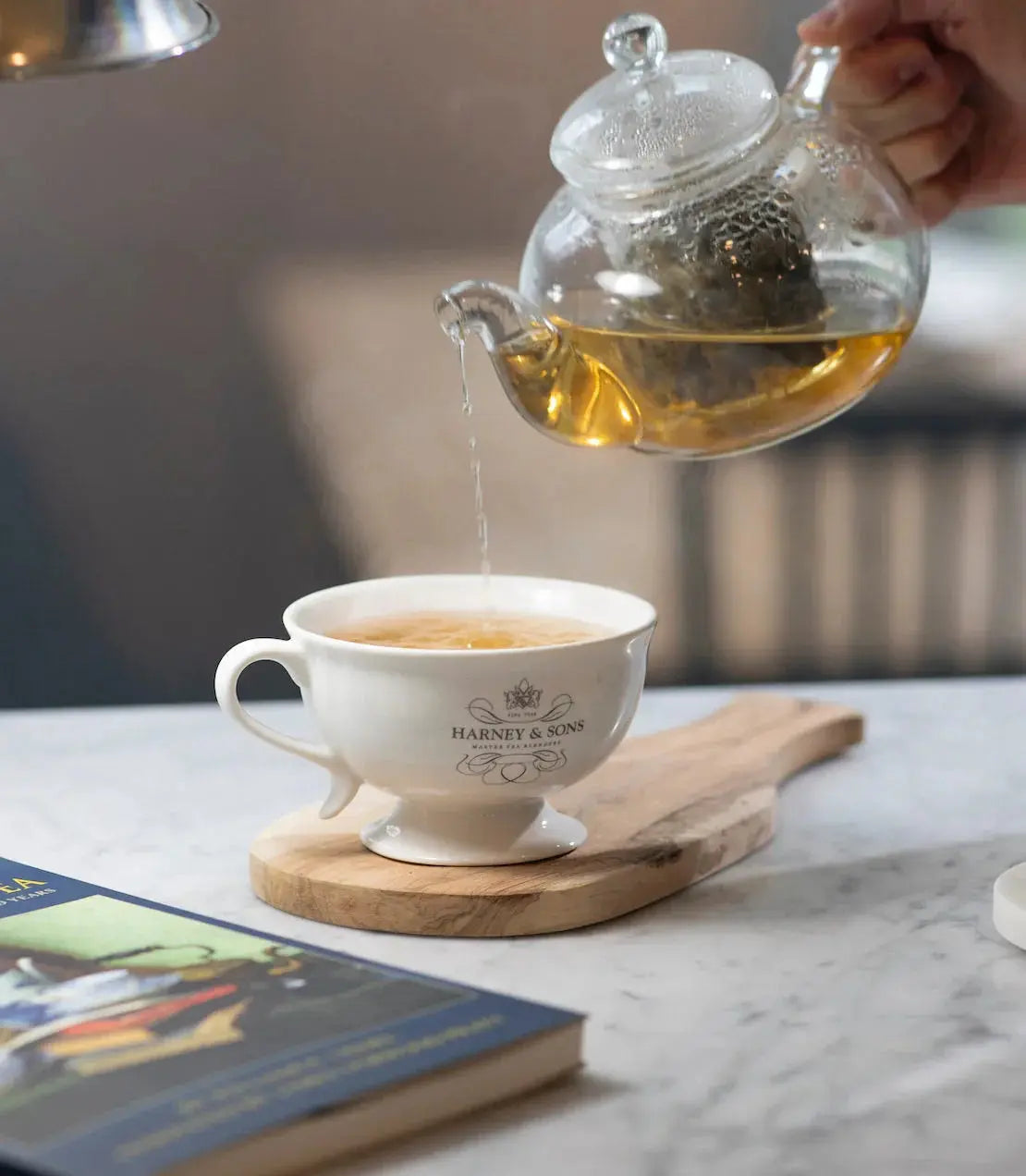 Pouring tea from glass teapot into Harney & Sons cup on wooden coaster, cozy farmhouse decor