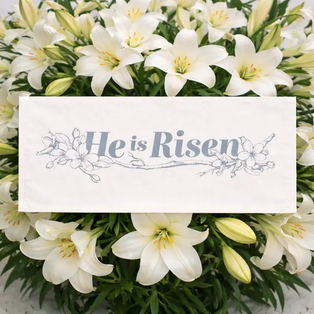 A white rectangular fabric banner or sign with the words “He is Risen” in blue script and floral line art, placed over a bouquet of white lilies.