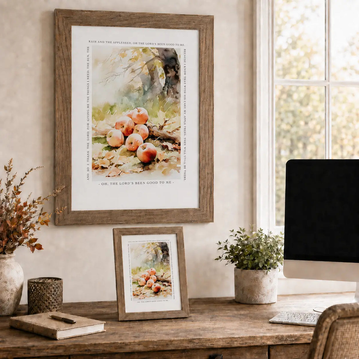 A framed art print featuring a watercolor still life of red apples on a desk in a warm, styled home office setting. A larger framed print hangs on the wall, with a smaller matching framed print resting on the wooden desk below, alongside a computer monitor, plants, and decorative items.
