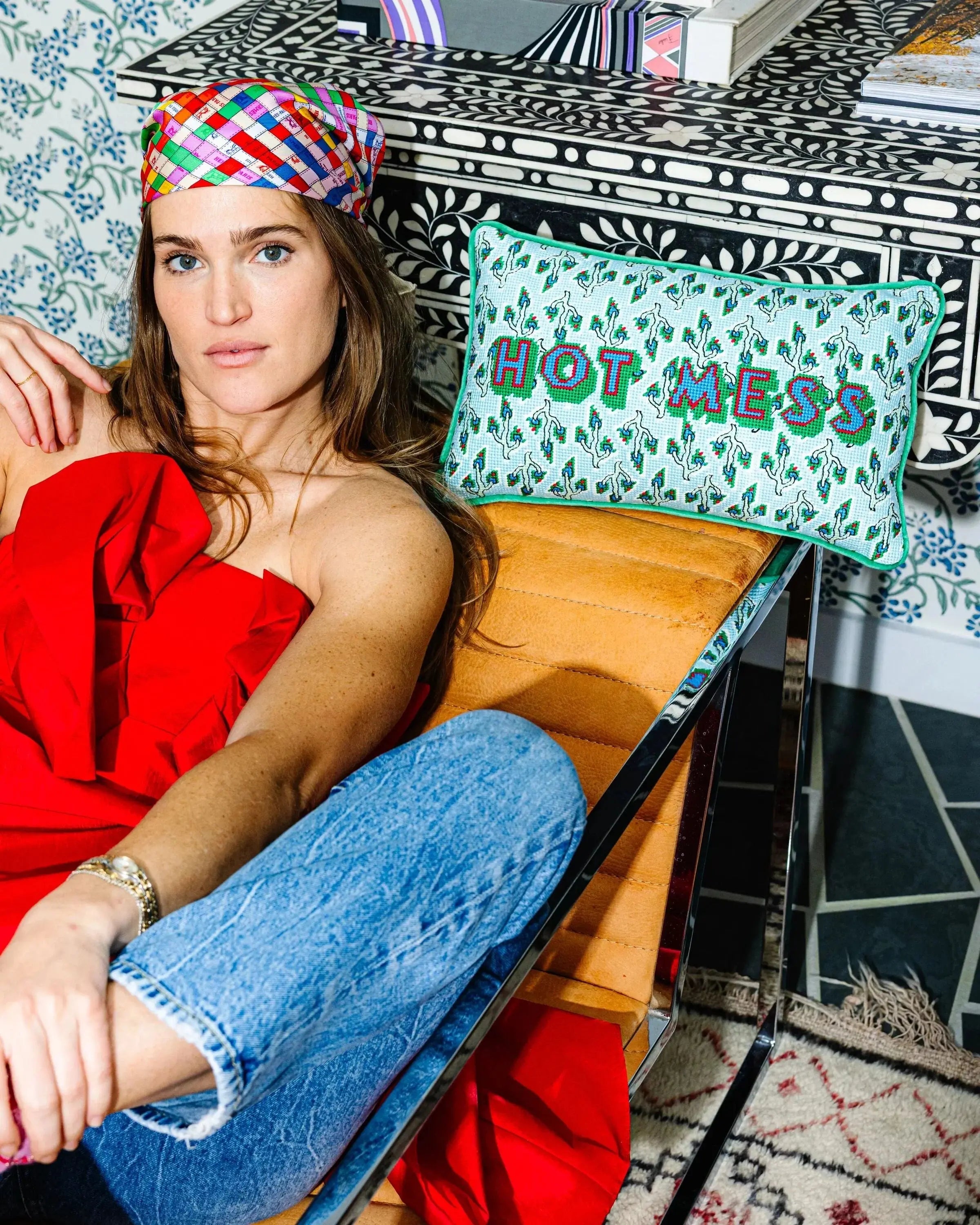 Woman in a red dress and colorful headscarf sitting on a chair next to a pillow with 'HOT MESS' text.