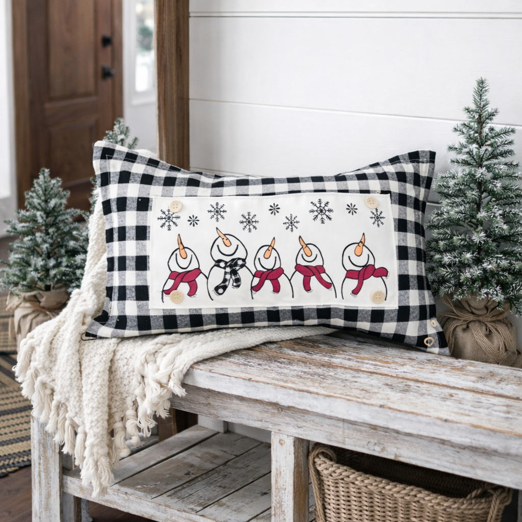 A rectangular throw pillow with a black and white buffalo check border and a central white panel featuring embroidered snowmen in red scarves and gray snowflakes, styled on a rustic wooden bench with a cream knit throw and small potted trees.