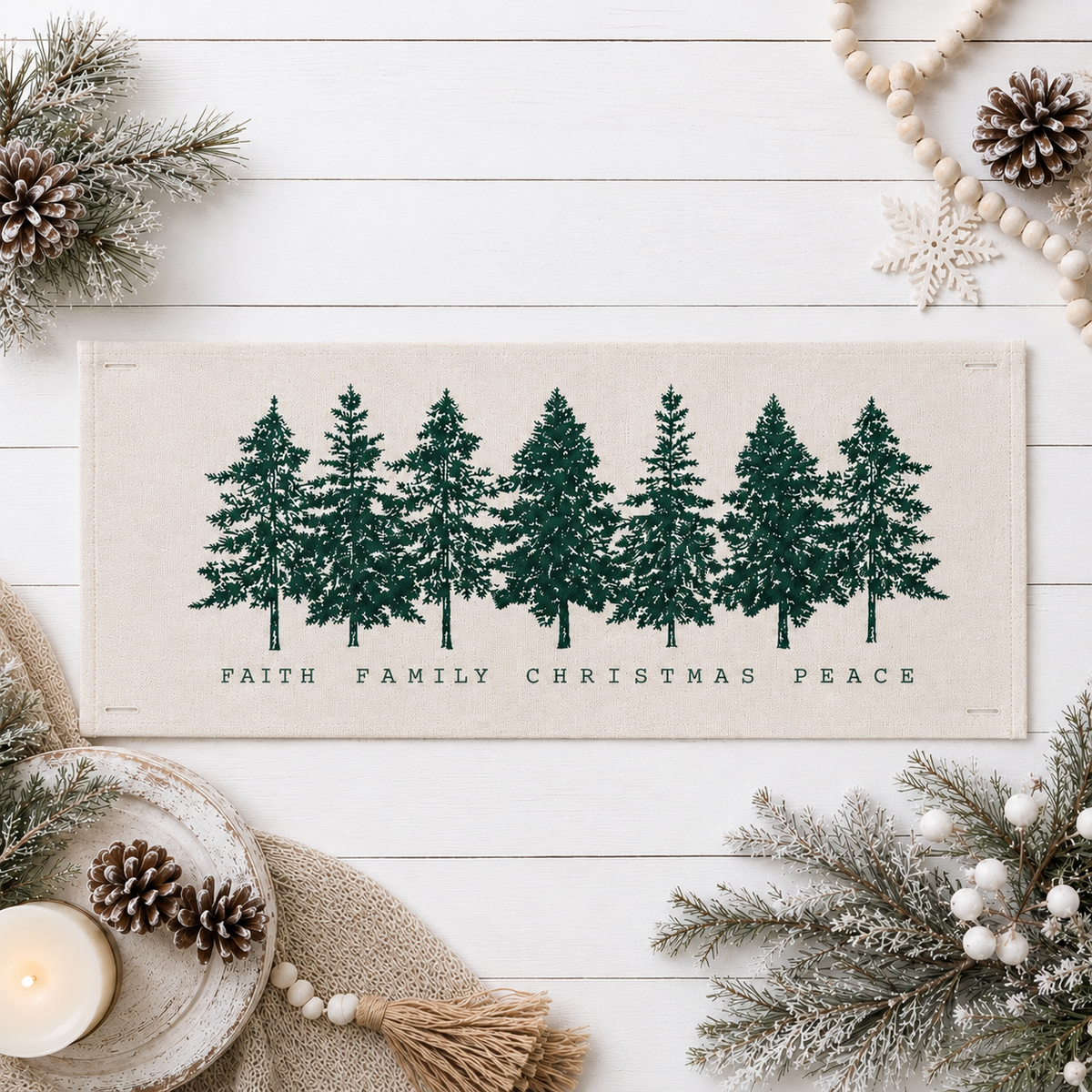 A rectangular beige fabric runner with a row of dark green pine tree silhouettes and the words 'FAITH FAMILY CHRISTMAS PEACE' printed below, styled on a white wood surface with holiday greenery, pinecones, beads, a candle, and a star ornament.
