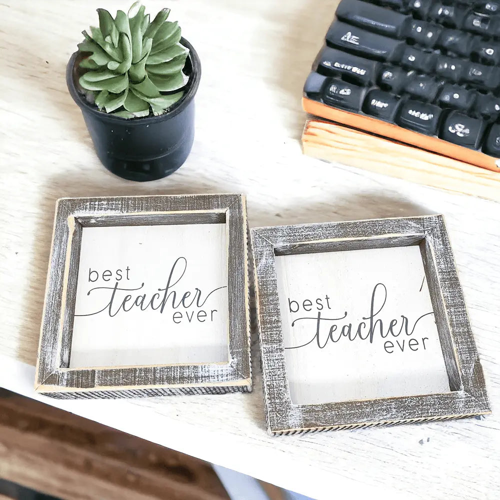 Two framed signs with 'best teacher ever' text on a desk with a keyboard and plant.