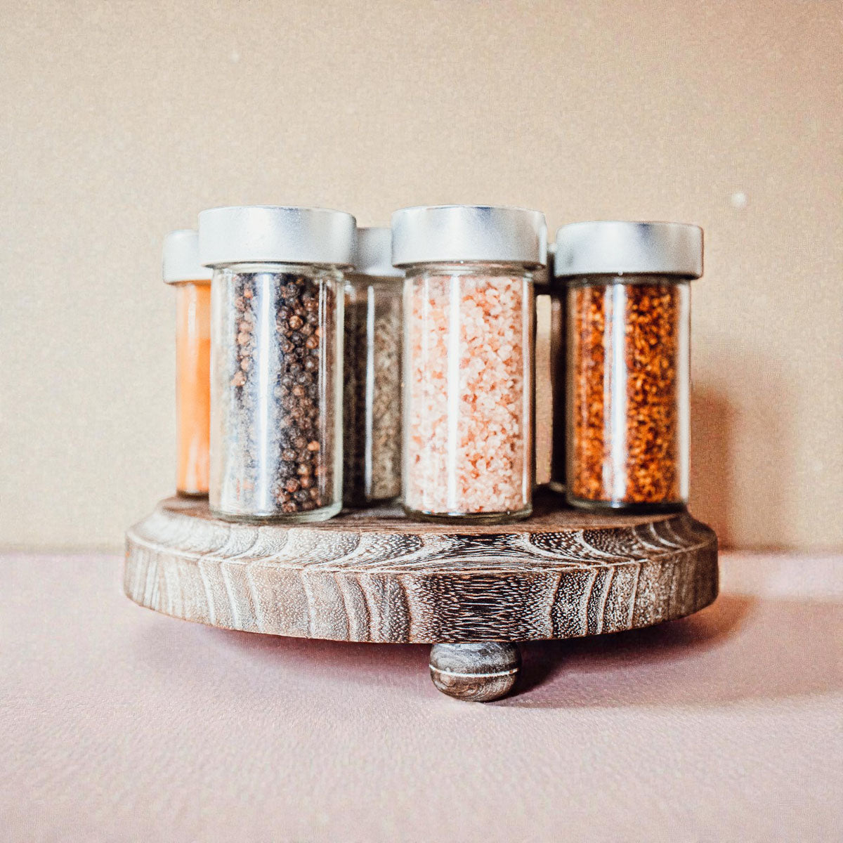 Set of five glass spice jars with metal lids on a wooden stand against a beige background