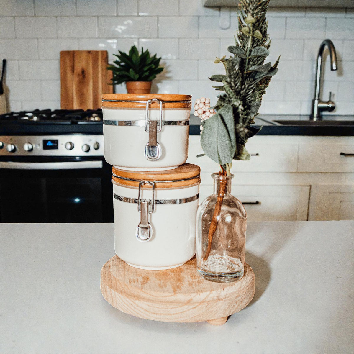 Two white canisters with wooden lids on a kitchen counter with a vase and plant.