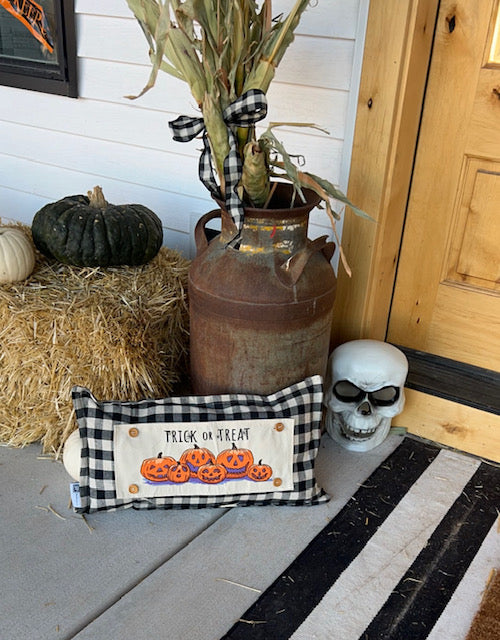 A rectangular black-and-white buffalo plaid pillow with embroidered orange pumpkins and the words 'TRICK OR TREAT' sits on a porch next to a rustic brown metal milk can, a hay bale with pumpkins, a dried corn bundle, and a white skull decoration.