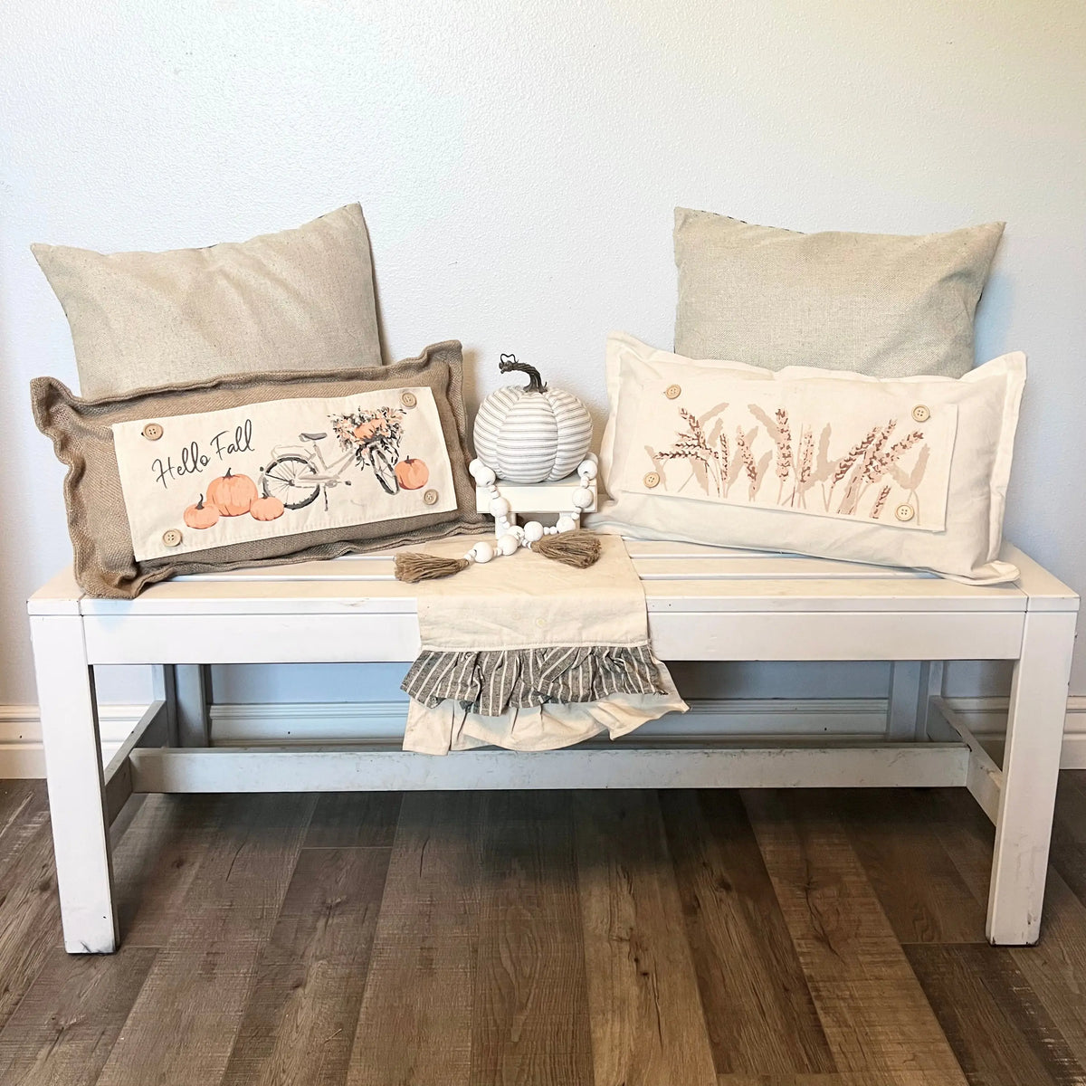 Two decorative throw pillows styled on a white wooden bench against a white wall, with a small striped pumpkin and a folded fringed throw blanket. The left pillow is brown with a 'Hello Fall' autumn design, and the right pillow is cream with a wheat design.