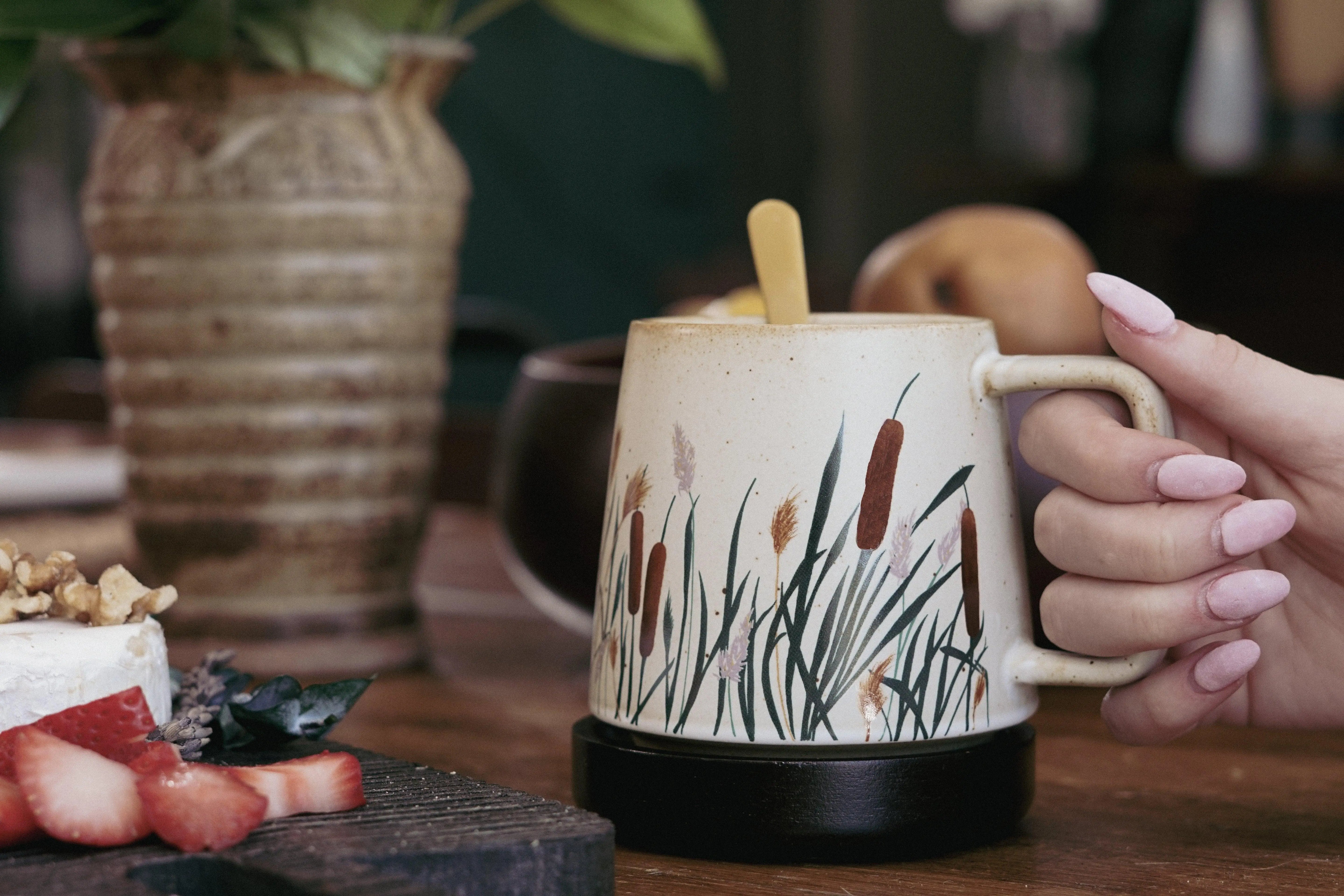 Hand holding rustic ceramic mug with cattail and wild grass design, farmhouse decor style